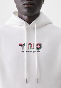 Hvit hettegenser med snor, prydet med flerfargede tekstlogoer "TRNO" og "the new originals" på forsiden. Myk tekstur.