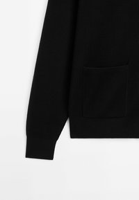 Manche et bas du torse d'un pull en maille côtelée noire avec une petite poche plaquée sur fond blanc.