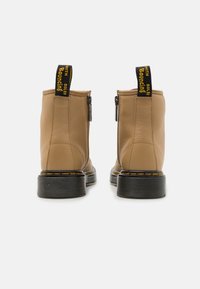Botas de cuero marrón hasta el tobillo con punta redonda, suela de goma negra, costuras amarillas y cremalleras laterales. Asa negra con texto amarillo en la parte trasera.