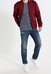 Viininpunainen bomber-takki valkoisen ja laivastonsinisen raitaisen paidan päällä, yhdistetty haalistuneisiin sinisiin farkkuihin ja valkoisiin lenkkareihin. Rentoa ja vartalonmyötäistä mallia.