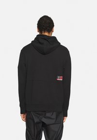 Hombre de espaldas vestido con sudadera negra con capucha caída y un parche en la parte baja de la espalda, combinado con pantalones negros holgados sobre fondo simple.