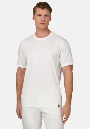 Homme aux cheveux courts et bouclés portant un t-shirt blanc uni à manches courtes et un pantalon blanc, debout devant un fond uni.