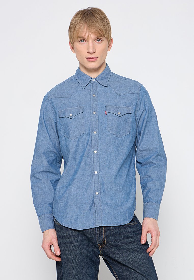 Camisa de mezclilla azul claro con mangas largas, cuello abotonado, dos bolsillos en el pecho y botones plateados, llevada con jeans oscuros.