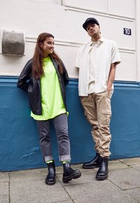 Giacca di pelle nera, maglietta verde neon, jeans grigi e stivali neri su una persona; un'altra in camicia a righe e pantaloni cargo color khaki. Sfondo di un muro blu.
