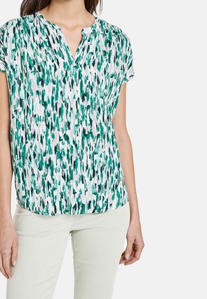 Femme portant un chemisier à manches courtes et col en V avec un imprimé abstrait en coups de pinceau verts et noirs, et un pantalon beige clair.
