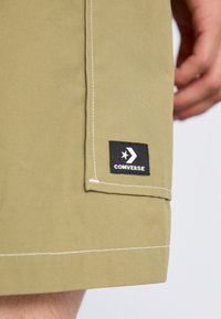Pantalones cortos verde oliva con corte recto, con costuras blancas y un parche del logo de Converse negro en el dobladillo. Textura de tela suave.