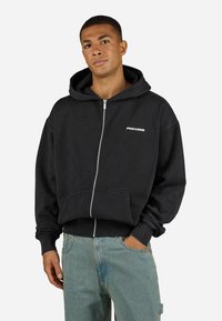 Black zip-up hoodie made of cotton with a front pocket, featuring a white "PEGADOR" logo on the left chest area. Ribbed cuffs and hem.