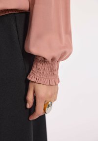 Różowa bluzka ze zgromadzonym mankietem, w połączeniu z czarnym materiałem. Ręka nosząca dużą srebrno-złotą pierścionek. Gładka faktura i luźny krój.