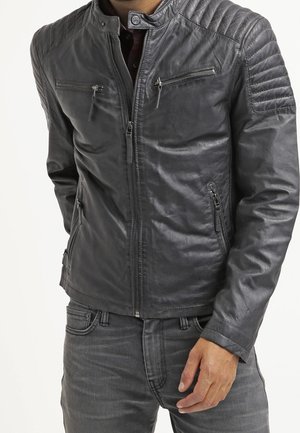 Homme portant une veste en cuir noire ajustée avec des détails matelassés aux épaules et des poches zippées, associée à un jean gris foncé.