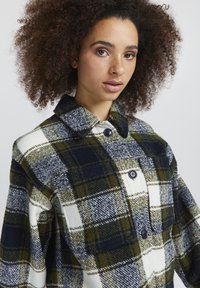 Plaid wool jacket featuring green, navy, and white colors, a collared neckline, large buttons, and a front pocket detail.