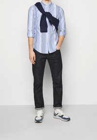 Man som bär uppvikta mörka jeans, vita och blå sneakers, en ljusblå randig skjorta med knappar och en marinblå tröja som hänger över axlarna.