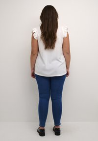 Weißes, kurzärmliges Oberteil mit Rüschendetails, kombiniert mit blauen engen Jeans und schwarzen Schuhen, von hinten betrachtet. Einfaches Design und Passform.