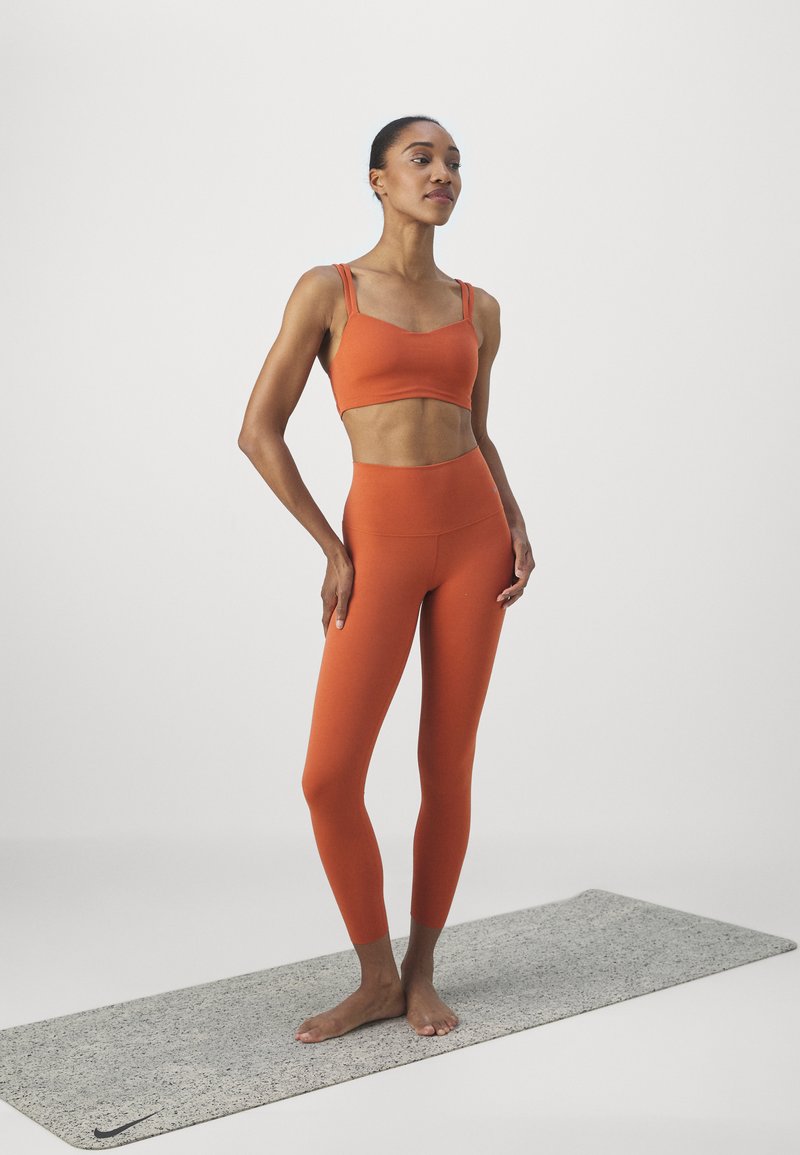 Reggiseno sportivo arancione e leggings a vita alta abbinati, texture liscia, design minimale, indossati su un tappetino da yoga grigio.