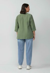 Groene, textuurbedekte shirt met een relaxte fit en driekwartmouwen, gecombineerd met lichtblauwe denim jeans en sneakers. Eentonige achtergrond.