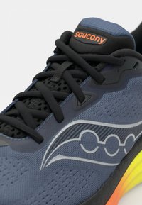 Zapatilla deportiva azul con parte superior de malla, cordones negros y logo lateral en plata. Presenta una suela texturizada con un degradado de amarillo a naranja.
