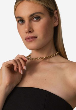 Gold chain necklace with rounded links, reflecting light, worn around the neck. Model holds the necklace, showing a black strapless top.