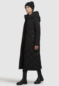 Abrigo largo negro acolchado con capucha, con costuras diagonales, bolsillos con cremallera y cierre frontal con cremallera. Combinado con un gorro de punto negro y botas.