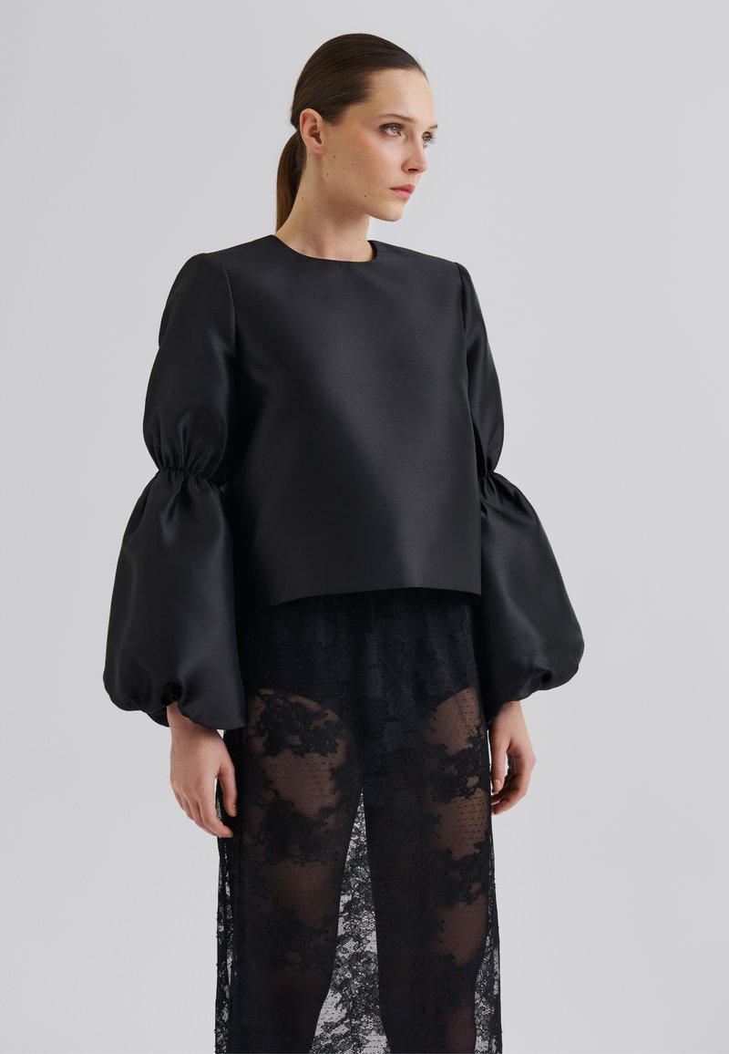 Svart cropped topp med pufferm, laget av glatt stoff, lagdelt over en gjennomsiktig blondeskjørt med blomstermønster og spaltedesign.