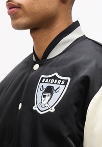 Κοντινό πλάνο ενός ατόμου που φοράει μαύρο και άσπρο μπουφάν Raiders με κουμπιά και το λογότυπο της ομάδας στο στήθος.