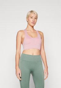 Brassière de sport rose cropped en tissu lisse, associée à des leggings verts taille haute en tissu côtelé. Design simple sans motifs ni accessoires.