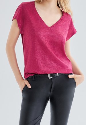 Kobieta ubrana w ciemnoróżową bluzkę z krótkim rękawem i dekoltem w serek, włożoną do czarnych dopasowanych spodni z czarnym paskiem, ręce w kieszeniach.