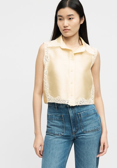 Νεαρή γυναίκα που φοράει αμάνικο κρεμ πουκάμισο με δαντέλα και ψηλόμεσο μπλε τζιν με μεγάλες μπροστινές τσέπες, κοιτάζοντας ελαφρώς προς τα πλάγια.