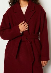 Femme portant un long manteau en laine bordeaux avec de larges revers et une ceinture nouée, main tenant l'avant du manteau près de la poitrine.