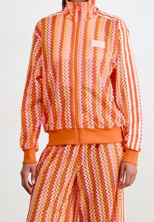 Femme portant une veste zippée et un pantalon assortis à motif zigzag orange, rose et jaune avec des poignets orange et des manches rayées.