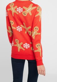 Pull rouge avec des bonshommes en pain d'épice et des flocons de neige en beige et blanc. Texture tricotée, poignets et ourlet côtelés, coupe décontractée.