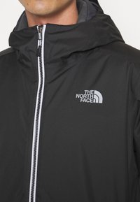 Veste à capuche noire avec une fermeture éclair à l'avant, ornée de accents blancs et d'une doublure intérieure grise. Le logo The North Face est affiché sur la poitrine.