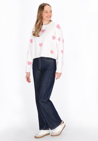 Pulover alb scurt cu modele de inimi roz, asortat cu blugi negri cu picior larg și adidași albi cu tălpi bej. Textură netedă.