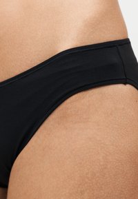 Gros plan d’une hanche portant un bas de bikini noir montrant une texture de peau lisse et les détails des coutures du vêtement.