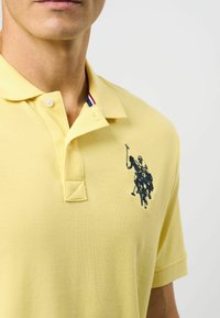 Žltá polo košeľa vyrobená z textúrovanej tkaniny, so zapínaním na tri gombíky a tmavomodrým vyšitým logom dvoch jazdcov na polo.