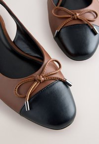 Ballerines marron avec bouts arrondis noirs, chacune ornée d'un nœud marron décoratif avec embouts métalliques sur un fond clair.
