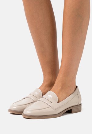 Mocasines de charol beige con punta redondeada, que presentan un detalle de doble tira y un tacón bajo y apilado. Textura suave y brillo sutil.