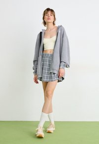 Szürke túlméretezett kapucnis pulóver, világos sárga crop top, szürke és fehér kockás szoknya, bézs sportcipő, fehér rövid zokni, zöld padlón áll.