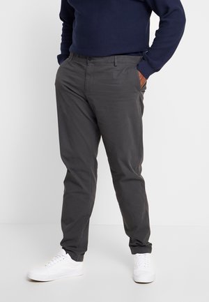 Mężczyzna ubrany w ciemnoszare spodnie, granatowy sweter i białe sneakersy, stoi z rękami w kieszeniach na jednolitym tle.