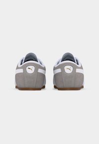 Ζευγάρι γκρι και λευκών αθλητικών παπουτσιών Puma, εμφανιζόμενα από πίσω, με χρυσά λογότυπα Puma στις φτέρνες και καφέ σόλες.