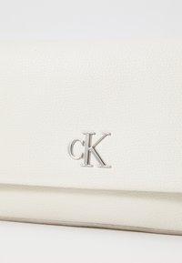 Balta odinė rankinė su tekstūruotu paviršiumi, sidabro spalvos detalėmis ir iškilniu "cK" logotipu, esančiu centre priekyje.