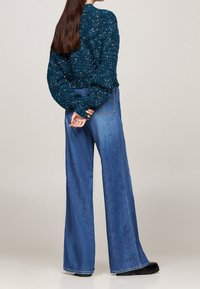 Vrouw met lang bruin haar, gekleed in een teal gebreid sweater en wijdvallende blauwe jeans, staand met de handen samengevoegd achter de rug.
