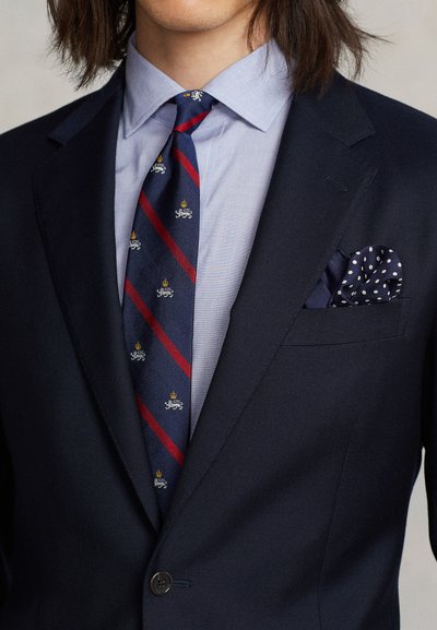 Marineblå dressjakke over en lyseblå skjorte, med et stripete slips med gule og røde innslag. Lommetørkle i marineblått med hvite prikker.