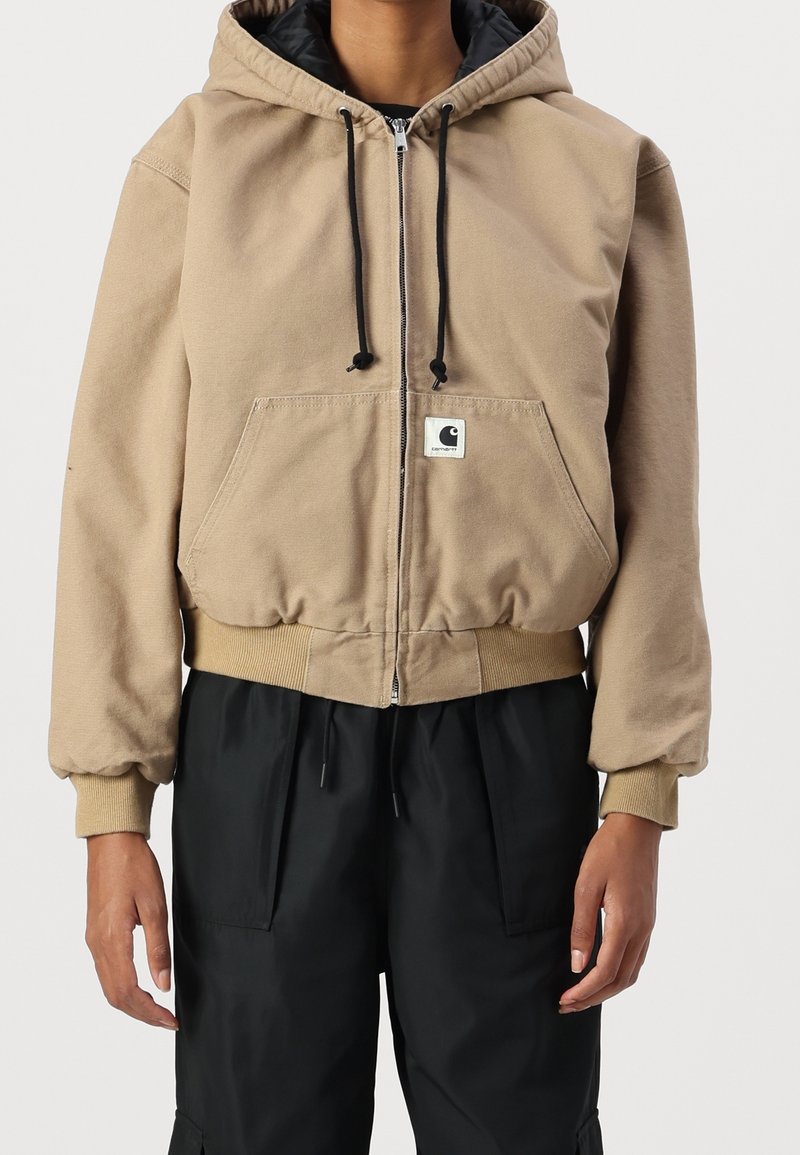 Beige zip-up hoodie met grote voorzak, trekkoorden capuchon en ribgebreide manchetten en onderkant. Gedragen over zwarte broek.