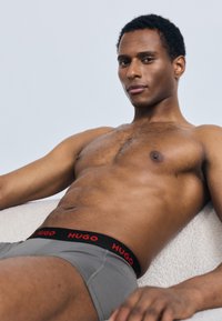 Gray boxer briefs with a black waistband featuring red "HUGO" branding. The model has dark skin and short curly hair, reclining on a light surface.
