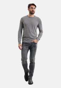 Maglione grigio a maglia con scollo rotondo, abbinato a jeans grigio scuro. Modello che indossa stivaletti neri in pelle, in piedi davanti a uno sfondo bianco.