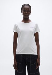 Fehér pamut póló kerek nyakkivágással és rövid ujjakkal, bal oldali mellrészen kis lila hímzett logóval.