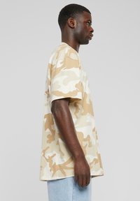 Ung mand iført en beige og hvid camouflage t-shirt og lyseblå jeans, der står i profil mod en ensfarvet grå baggrund.