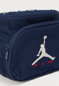 Τσάντα από υφασμάτινο σκούρο μπλε ύφασμα με εμπρόσθια τσέπη με φερμουάρ, που διαθέτει λευκό λογότυπο Jumpman και κόκκινο κείμενο "AIR". Υφή με ορατές ραφές.