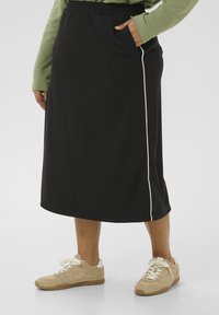 Jupe midi noire avec une taille élastique et des passepoils blancs sur les côtés. Associée à des baskets beige clair avec une texture en daim et des accents blancs.