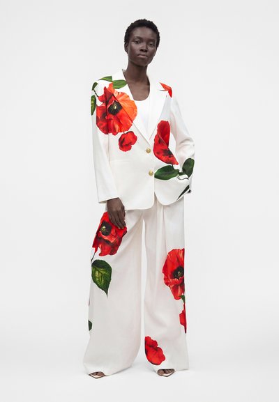 Femme portant un costume blanc avec de grandes fleurs de coquelicot rouges imprimées, un pantalon à jambes larges et un blazer à double boutonnage, debout sur un fond blanc.