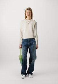Off-white ribbed sweater, relaxed fit, blue distressed jeans, green gradient mesh bag, white sneakers, natural hair. Plain background.